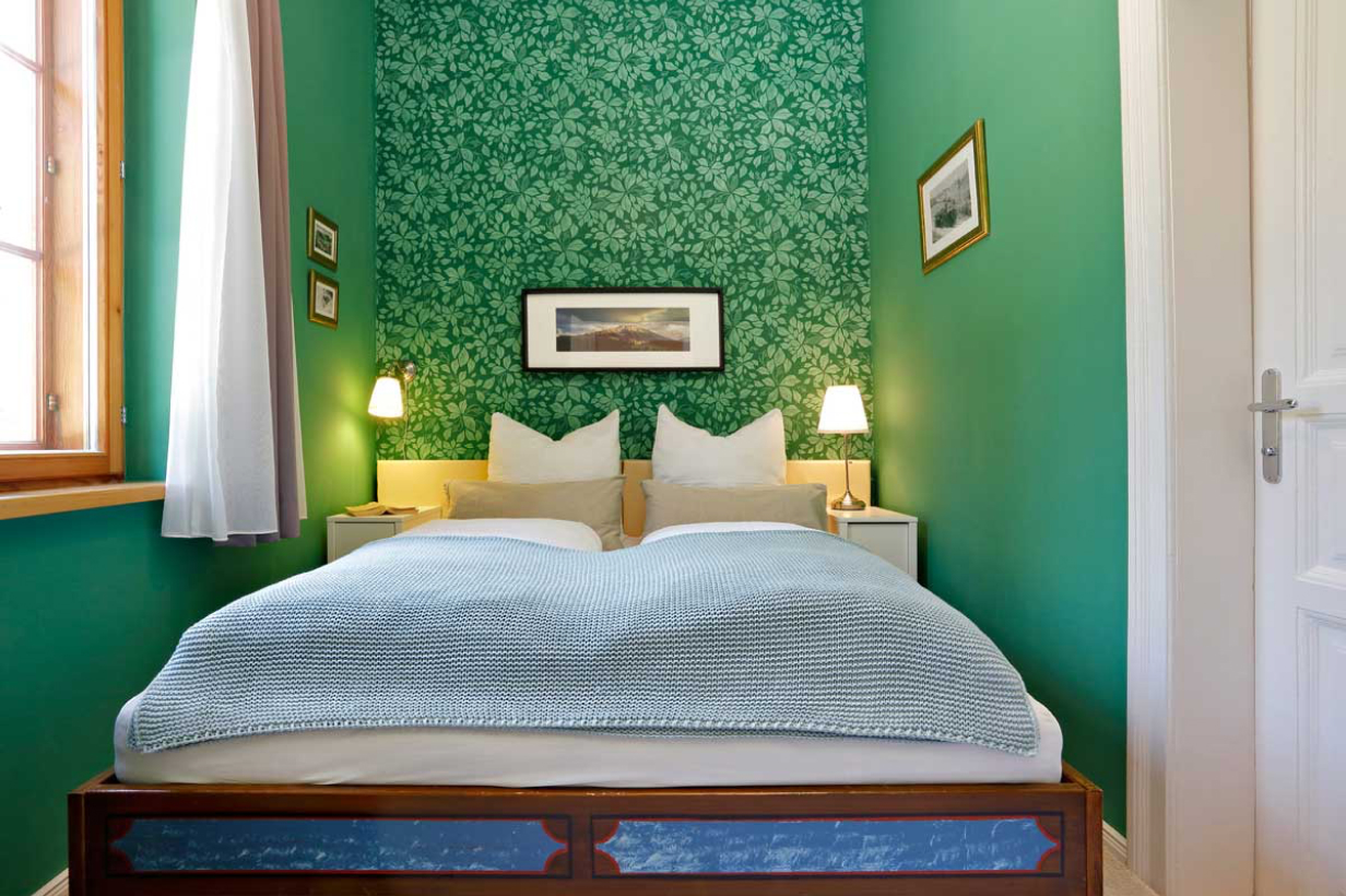 Ein gemütliches Schlafzimmer mit grünen Wänden, einer gemusterten grünen Akzentwand, einem Doppelbett mit weißer und grauer Bettwäsche, zwei Nachttischen mit Lampen, gerahmten Bildern an den Wänden und einem Fenster mit durchsichtigen Vorhängen.