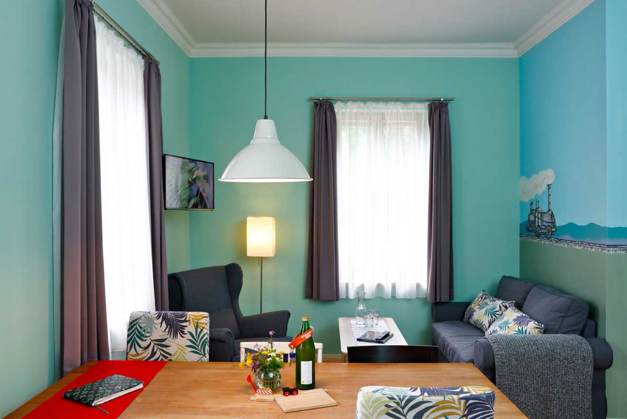 Ein gemütliches, helles Wohnzimmer mit türkisfarbenen Wänden, einem Holztisch für zwei Personen, grauen Sesseln, einem grauen Sofa und großen Fenstern mit durchsichtigen Vorhängen. Es gibt eine moderne Einrichtung, eine Hängelampe und ein Gemälde an der Wand.