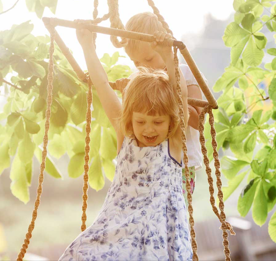 urlaub-am-bahnhof-garten-ideal-fuer-kinder Zwei kleine Kinder spielen auf einer Seilschaukel im Freien. Das Sonnenlicht fällt durch die grünen Blätter um sie herum. Ein Kind in einem geblümten Kleid hängt lächelnd an den Seilen, während das andere hinter ihm steht und hilft.