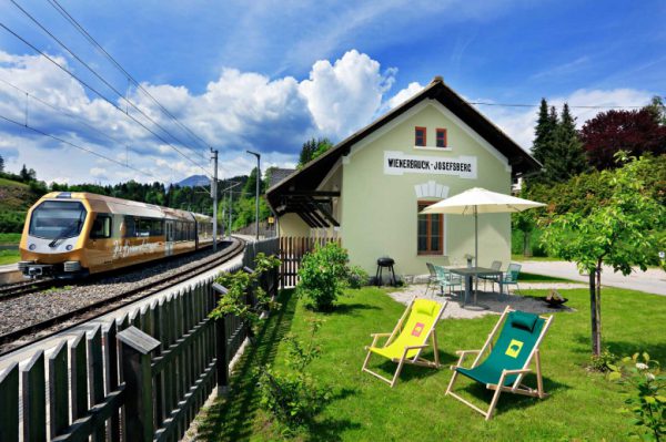 front-2 Ein gelber Zug fährt an einem kleinen Bahnhofsgebäude mit der Aufschrift Weißenbach-Josefsberg vorbei. Zwei Liegestühle und ein Tisch mit einem Sonnenschirm stehen auf dem grünen Rasen neben einem Holzzaun unter einem teilweise bewölkten Himmel.
