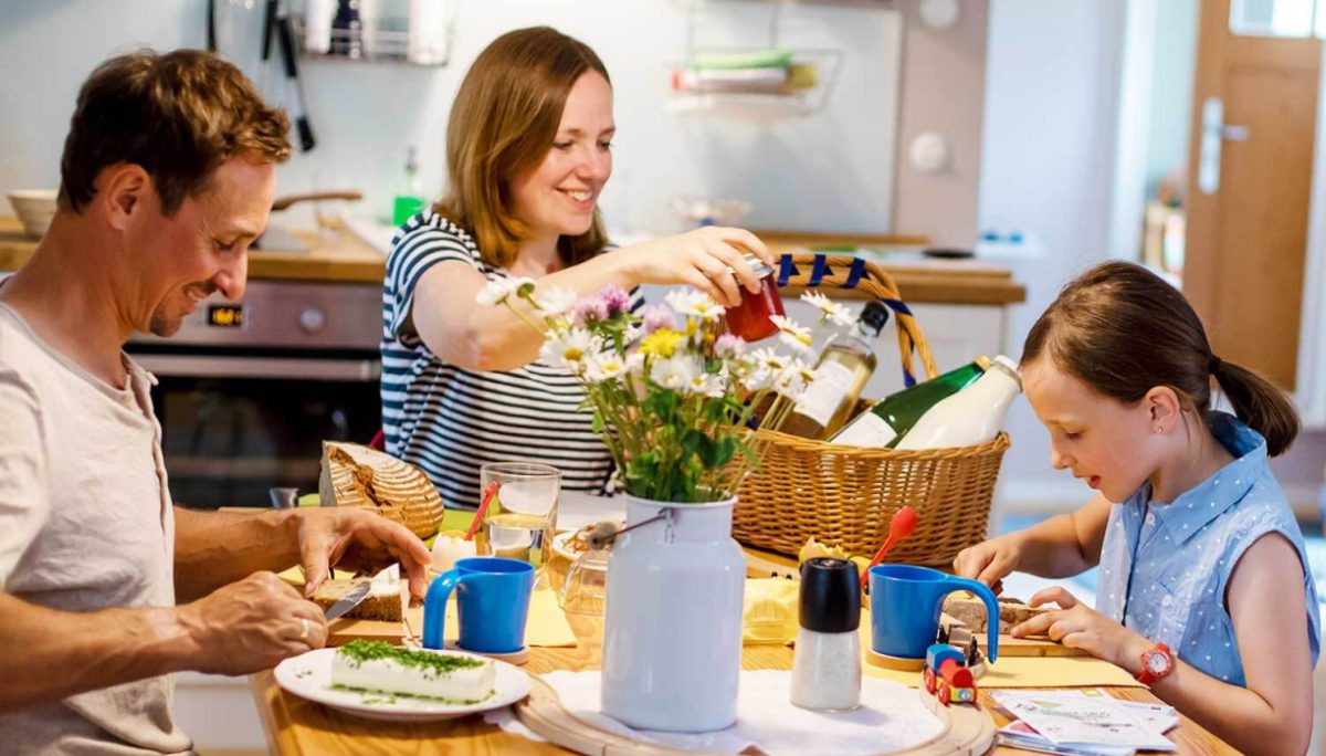 Eine dreiköpfige Familie sitzt an einem Küchentisch, lächelt und bereitet gemeinsam das Essen zu. Auf dem Tisch stehen Tassen, Brot und ein Korb mit Lebensmitteln. Die Umgebung ist hell und fröhlich, in der Mitte steht eine Vase mit Wildblumen.