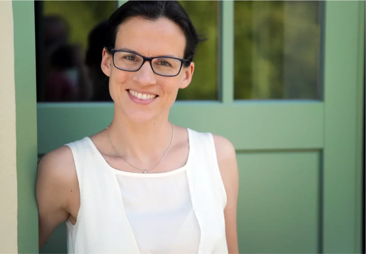 ueber-uns-2 Eine Frau mit dunklem Haar und Brille, die ein ärmelloses weißes Oberteil trägt, steht lächelnd vor einer grünen Tür mit Glasscheiben.