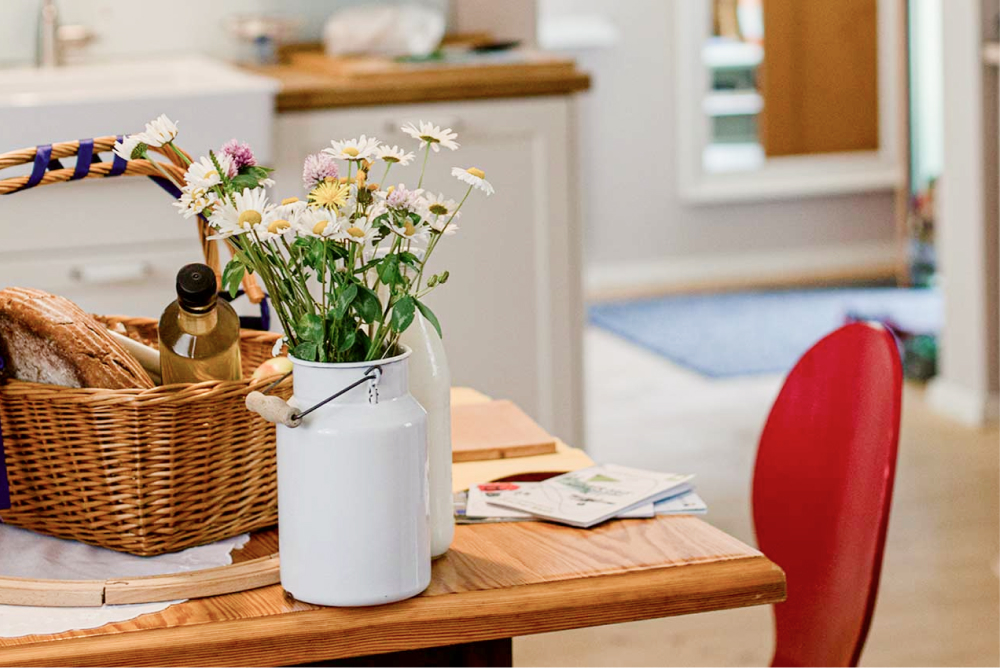 Ein Holztisch mit einem weißen Metallkrug mit Wildblumen, einem Weidenkorb mit Brot und einer Flasche sowie verstreuter Post; im Vordergrund ein roter Stuhl, im Hintergrund eine unscharfe Küchenzeile.