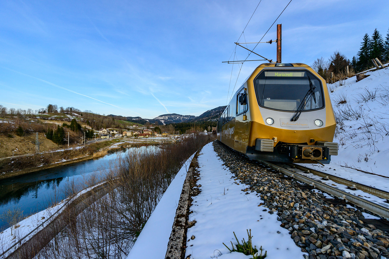 mariazellerbahn-wolfgang-stecher_WST7752 1 Ein moderner gelber Zug fährt auf einer verschneiten Eisenbahnstrecke neben einem Fluss, mit Hügeln, Häusern und Bäumen im Hintergrund unter blauem Himmel.