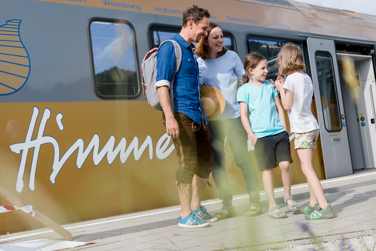 Eine vierköpfige Familie, zwei Erwachsene und zwei Kinder, steht lächelnd und plaudernd auf einem Bahnsteig neben einem goldfarbenen Zug mit großen weißen Buchstaben an der Seite. Das Wetter ist sonnig und hell.