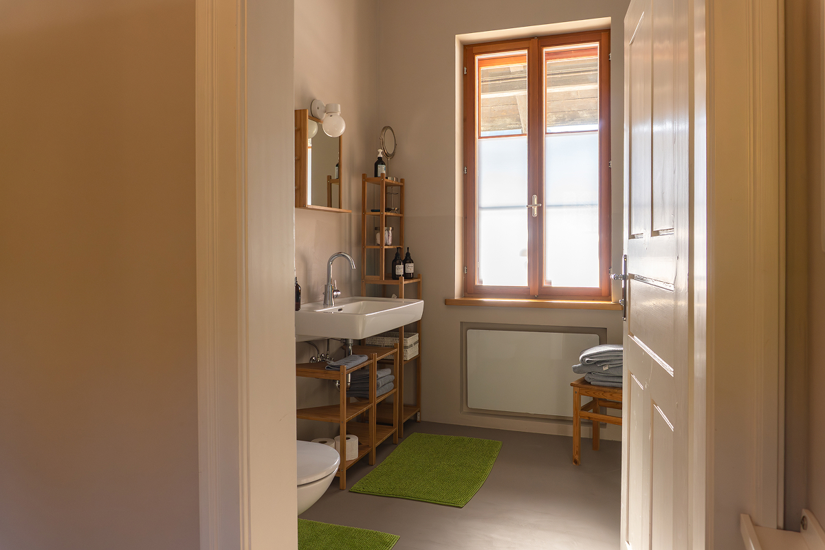 Ein helles, aufgeräumtes Badezimmer mit einem großen Fenster, einem weißen Waschbecken, Holzregalen mit Handtüchern und Toilettenartikeln, grünen Fußmatten und neutralen Wänden, von der offenen Tür aus gesehen.