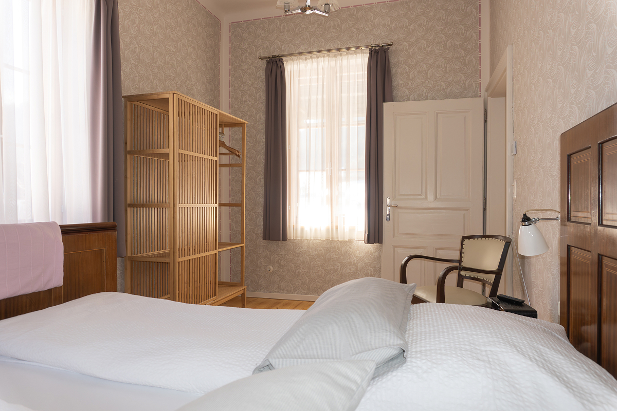 Ein helles, aufgeräumtes Schlafzimmer mit einem gemachten Bett, einem Holzschrank, einem Sessel, einer Lampe und hellen Vorhängen, die das Sonnenlicht durch die Fenster hereinlassen und eine warme und einladende Atmosphäre schaffen.