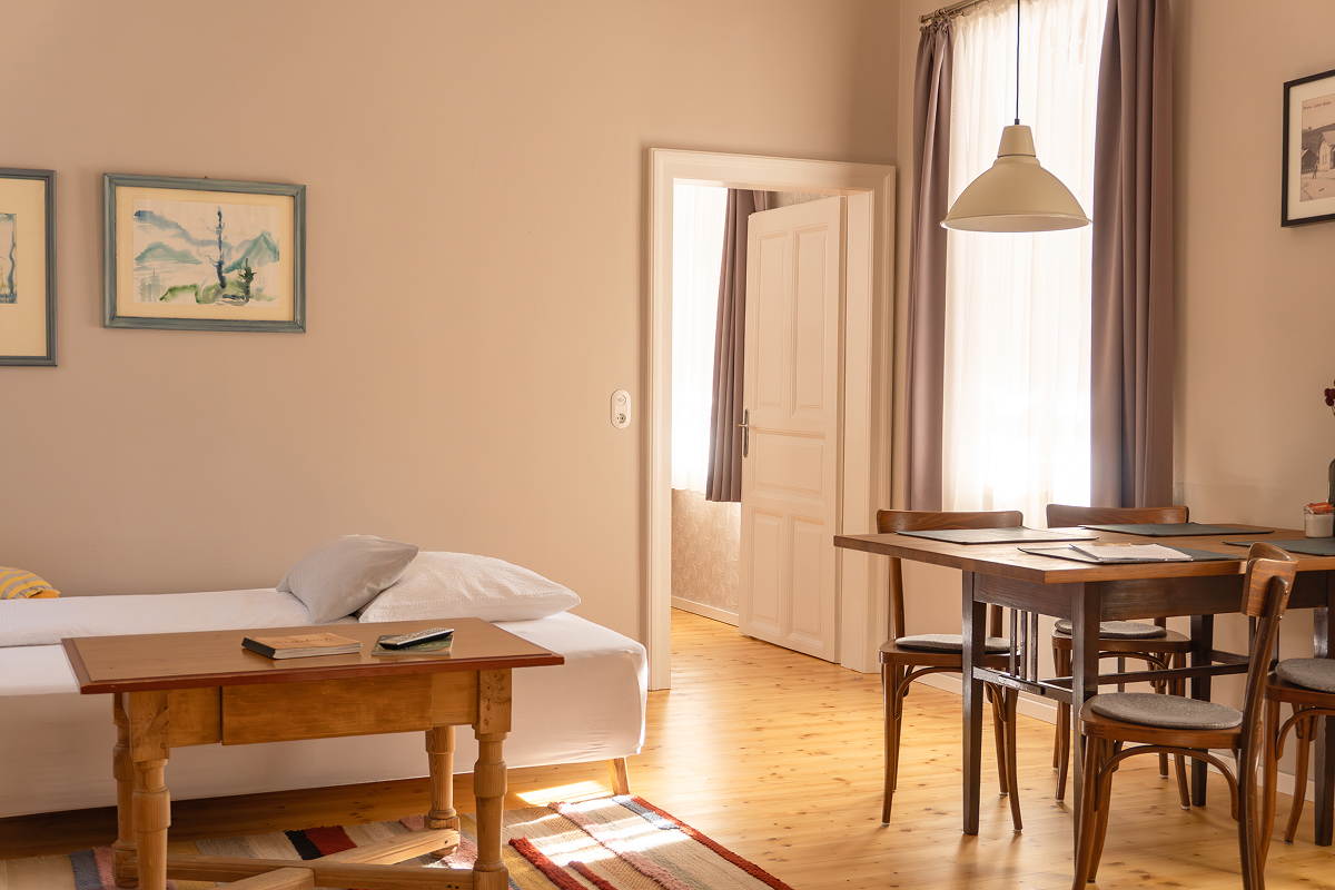 Ein gemütliches, sonnendurchflutetes Zimmer mit einem Einzelbett, einem Holztisch, Stühlen, Wandkunst und großen Fenstern mit Vorhängen. Das Licht strömt durch eine offene Tür herein und schafft eine warme, einladende Atmosphäre.