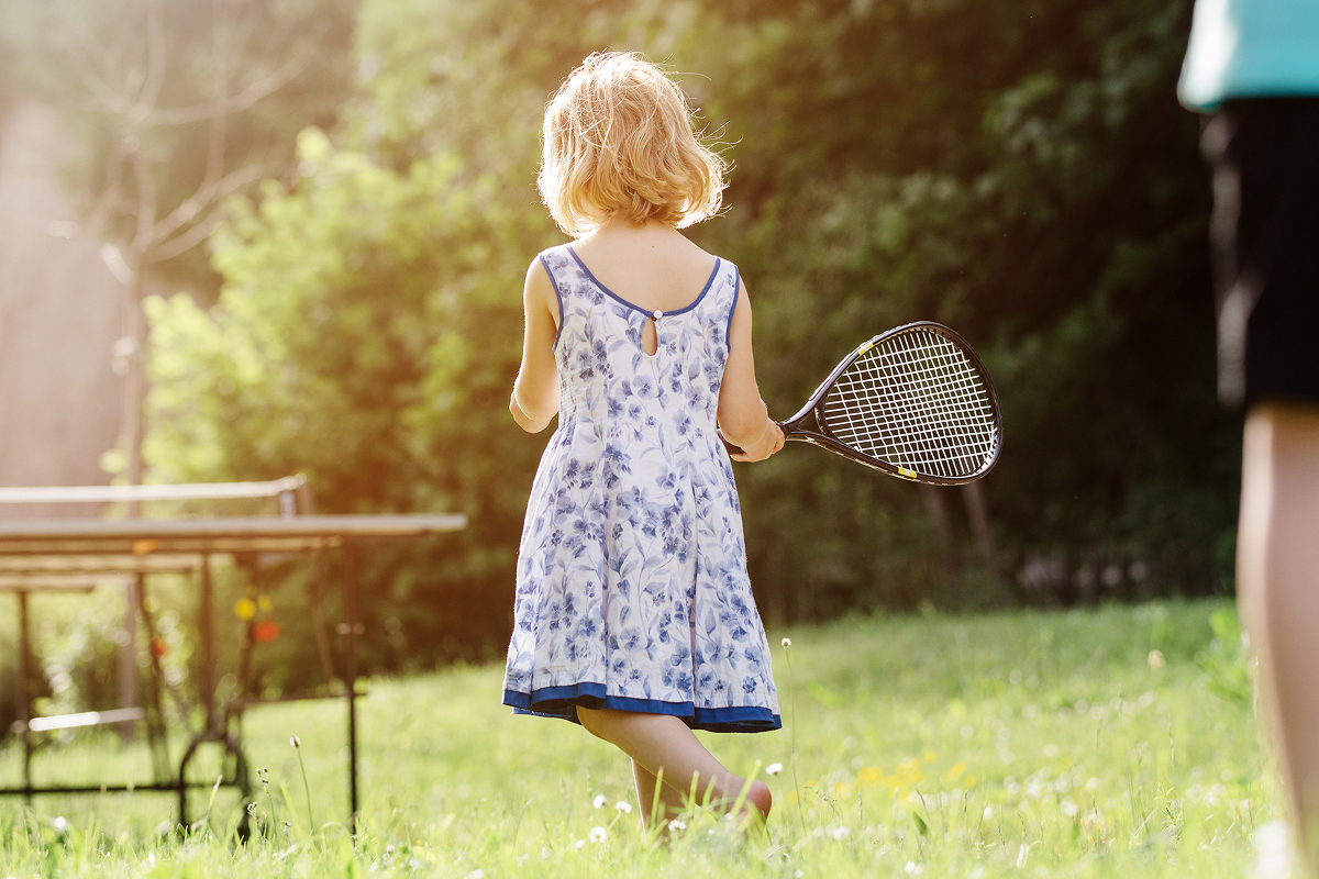 Ein junges Mädchen in einem blau-weißen Kleid hält einen Tennisschläger in der Hand, während es auf einer Wiese neben einer Tischtennisplatte steht, mit Bäumen und Sonnenlicht im Hintergrund.