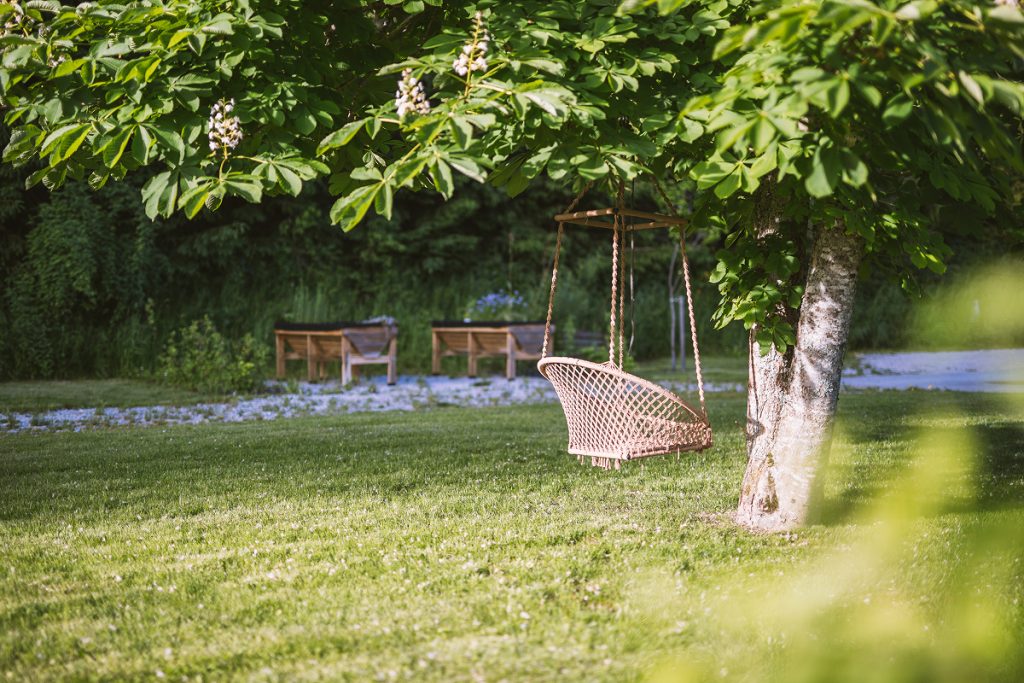 Eine Korbschaukel hängt an einem belaubten Baum in einem sonnenbeschienenen, grasbewachsenen Garten, mit zwei Holzstühlen und einem kleinen Tisch im Hintergrund neben einem Kiesweg.