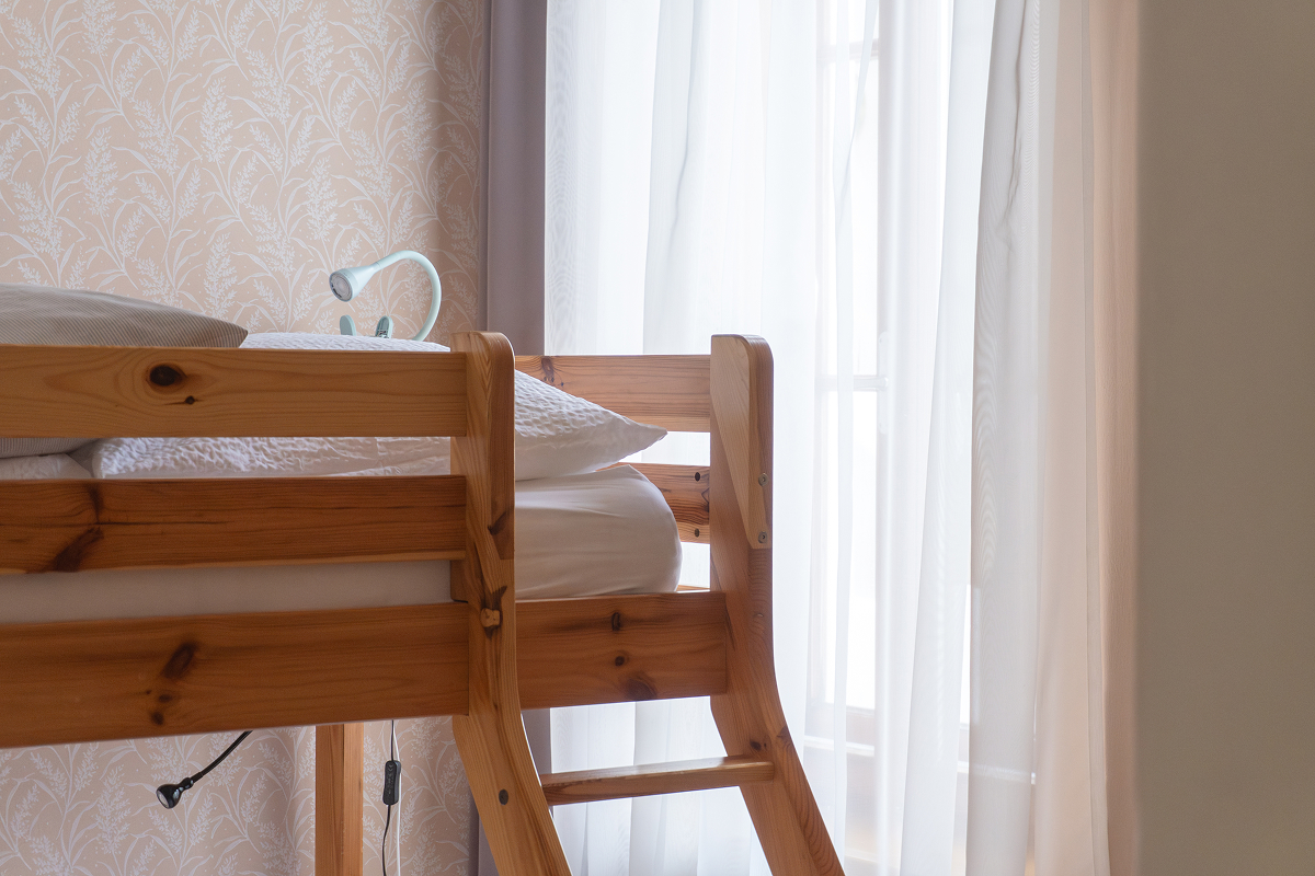Ein hölzernes Etagenbett mit weißem Bettzeug steht neben einem Fenster mit weißen Vorhängen. Sanftes Tageslicht fällt in den gemütlichen Raum und hebt die gemusterte Tapete und eine kleine Leselampe am Bett hervor.