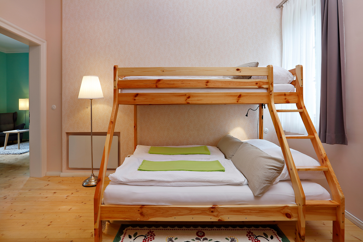 Ein gemütliches Schlafzimmer mit einem hölzernen Etagenbett mit einem Doppelbett unten und einem Einzelbett oben, sauber gemacht mit weißer Bettwäsche und grünen Handtüchern. Das Zimmer hat eine Stehlampe, einen hellen Holzfußboden und ein Fenster mit durchsichtigen Vorhängen.