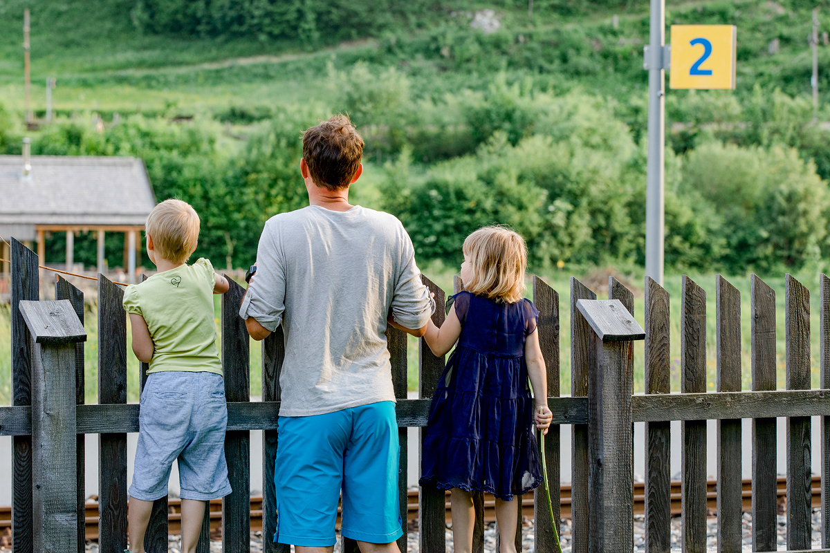 Ein Mann und zwei kleine Kinder stehen an einem Holzzaun und schauen auf die Bahngleise. Die Umgebung ist grün und üppig, im Hintergrund ist ein Schild mit der Nummer 2 zu sehen. Der Mann und die Kinder stehen mit dem Rücken zur Kamera.