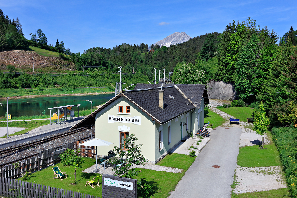 Ein kleines Bahnhofsgebäude mit der Aufschrift Weißenbach-Werfenbahn liegt neben den Bahngleisen, mit einem Fluss, Bäumen, grünen Hügeln und einem entfernten Berg im Hintergrund unter einem klaren blauen Himmel.