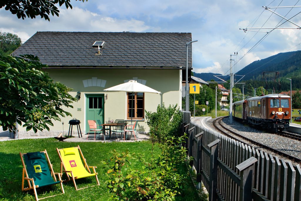 Ein Haus mit einem eingezäunten Hof, Terrassenmöbeln und zwei bunten Liegestühlen steht neben einer Eisenbahnstrecke, während sich ein alter Zug unter einem teilweise bewölkten Himmel nähert. Üppiges Grün umgibt die Szene.