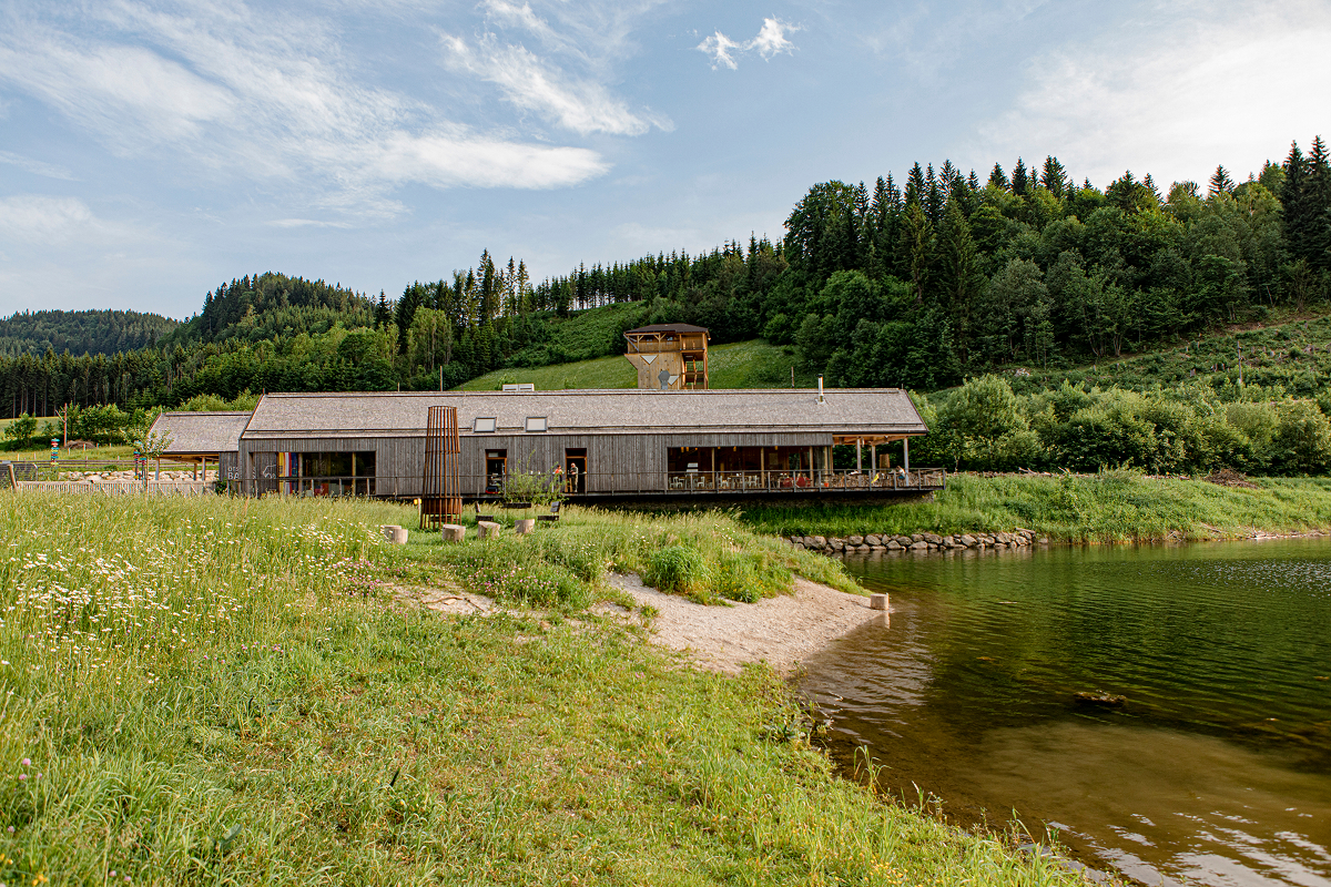 Ein Holzgebäude steht neben einem ruhigen Teich, umgeben von saftig grünem Gras und Wildblumen, mit bewaldeten Hügeln und einem teilweise bewölkten Himmel im Hintergrund.