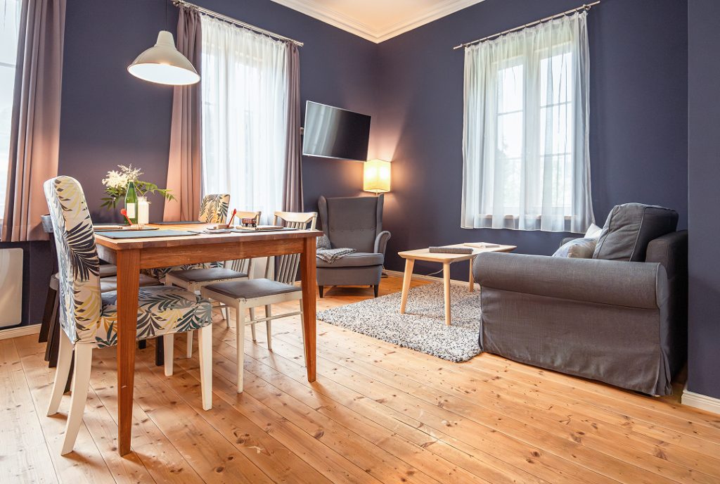 Ein gemütlicher Wohn- und Essbereich mit dunkelblauen Wänden, Holzboden, einem Esstisch mit verschiedenen Stühlen, einem grauen Sessel, einem grauen Sofa, einem Fernseher an der Wand und großen Fenstern mit weißen Vorhängen.