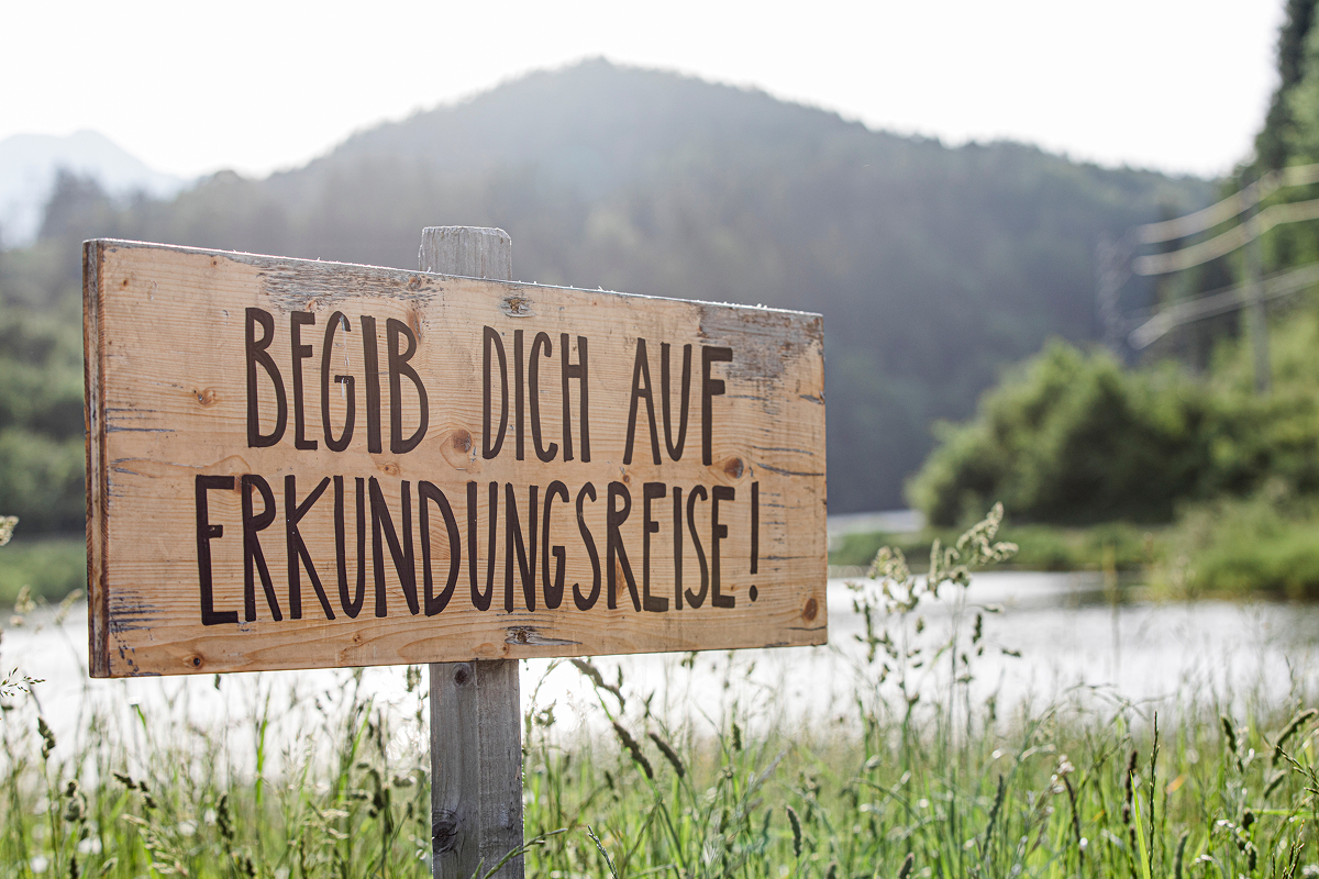 Auf einem hölzernen Schild auf einer Wiese steht "Begib dich auf Erkundungsreise!" auf Deutsch, mit einem See und bewaldeten Hügeln im Hintergrund unter einem klaren Himmel.