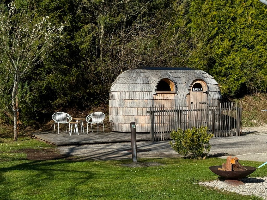 Hinter einem schwarzen Metallzaun steht eine kleine hölzerne Saunahütte mit einem runden Dach. Zwei weiße Stühle und ein Tisch stehen in der Nähe, umgeben von grünem Gras und Bäumen im Hintergrund.
