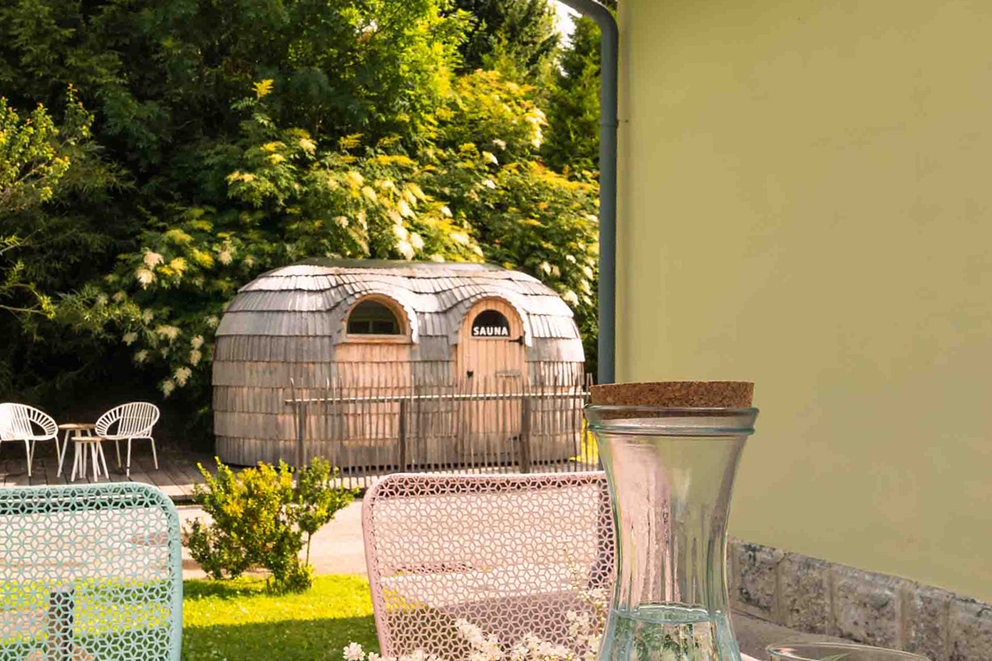 sauna-garten-ferienhaus-bahnhof-oetschergraeben-11 Eine kleine, rustikale Saunahütte aus Holz steht in einem sonnigen Garten, umgeben von Grünpflanzen. Im Vordergrund stehen pastellfarbene Stühle und eine Glaskaraffe auf einem Tisch. Im Hintergrund sind weiße Außenstühle zu sehen.