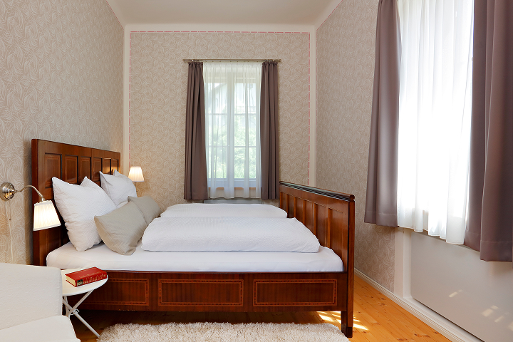 Ein gemütliches Schlafzimmer mit einem hölzernen Bettgestell, weißem Bettzeug und mehreren Kissen. Das Zimmer hat helle gemusterte Tapeten, ein Fenster mit taupefarbenen Vorhängen, eine kleine Nachttischlampe und einen weißen Stuhl mit einem roten Buch.