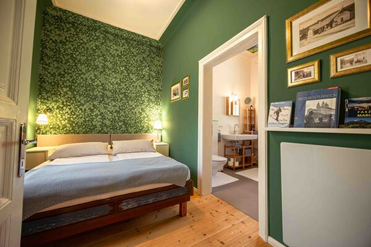Ein gemütliches Schlafzimmer mit grünen Wänden, einem Doppelbett, zwei Wandlampen und einer Blättertapete hinter dem Bett. Das Zimmer führt zu einem angrenzenden Badezimmer, und es gibt Bücher und gerahmte Bilder auf einem Regal und an der Wand.