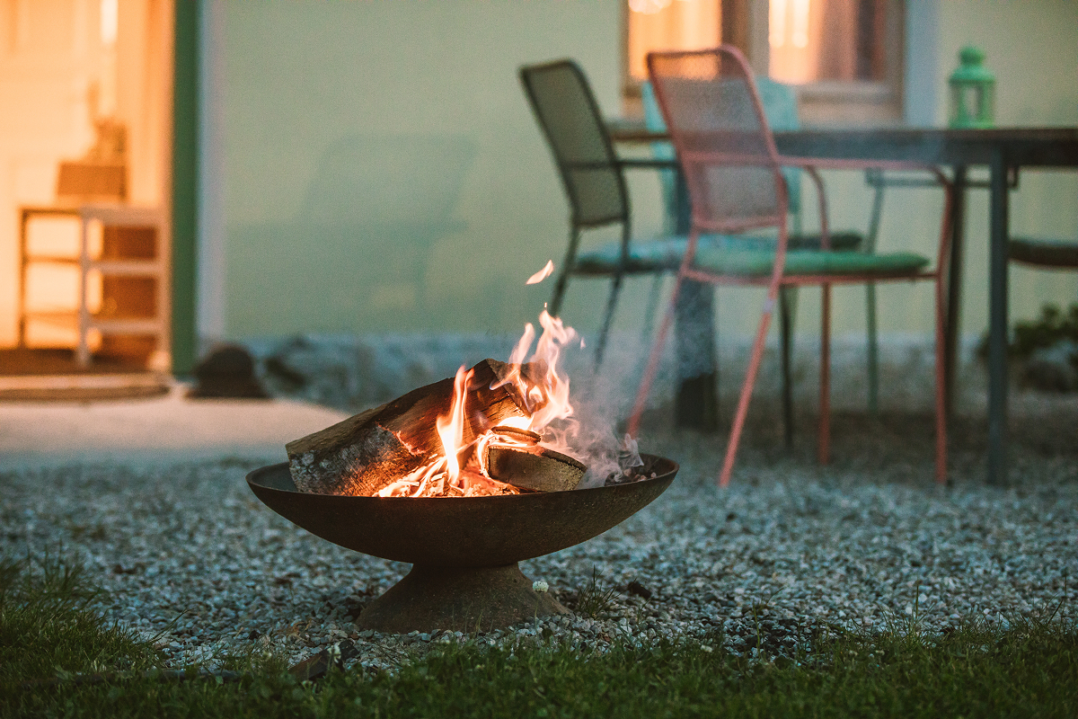Eine Feuerstelle aus Metall mit brennenden Holzscheiten steht auf einer Kiesfläche, daneben stehen Stühle und ein Tisch. Im Hintergrund ist ein Haus mit offener Tür und gedämpfter Innenbeleuchtung zu sehen.