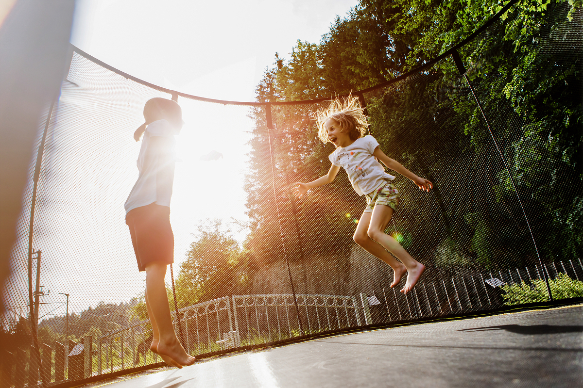 Zwei Kinder springen an einem sonnigen Tag im Freien auf einem Trampolin. Das Sonnenlicht fällt durch die Bäume im Hintergrund und schafft eine spielerische und energiegeladene Atmosphäre. Beide Kinder scheinen Spaß zu haben.