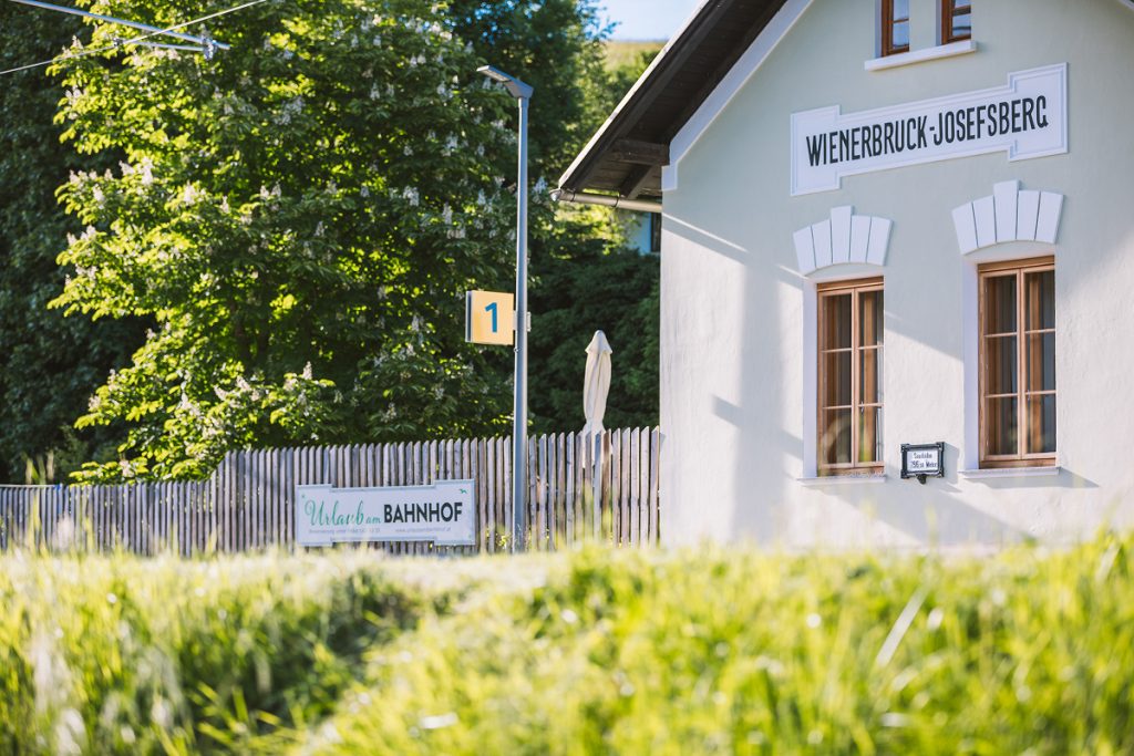 Ein kleines Bahnhofsgebäude mit der Aufschrift "Wienerbruck-Josefsberg" steht an einem Zaun, das Bahnsteigschild "1" und ein "Bahnhof"-Schild sind sichtbar. Üppiges grünes Gras und Bäume umgeben die Szene an einem sonnigen Tag.