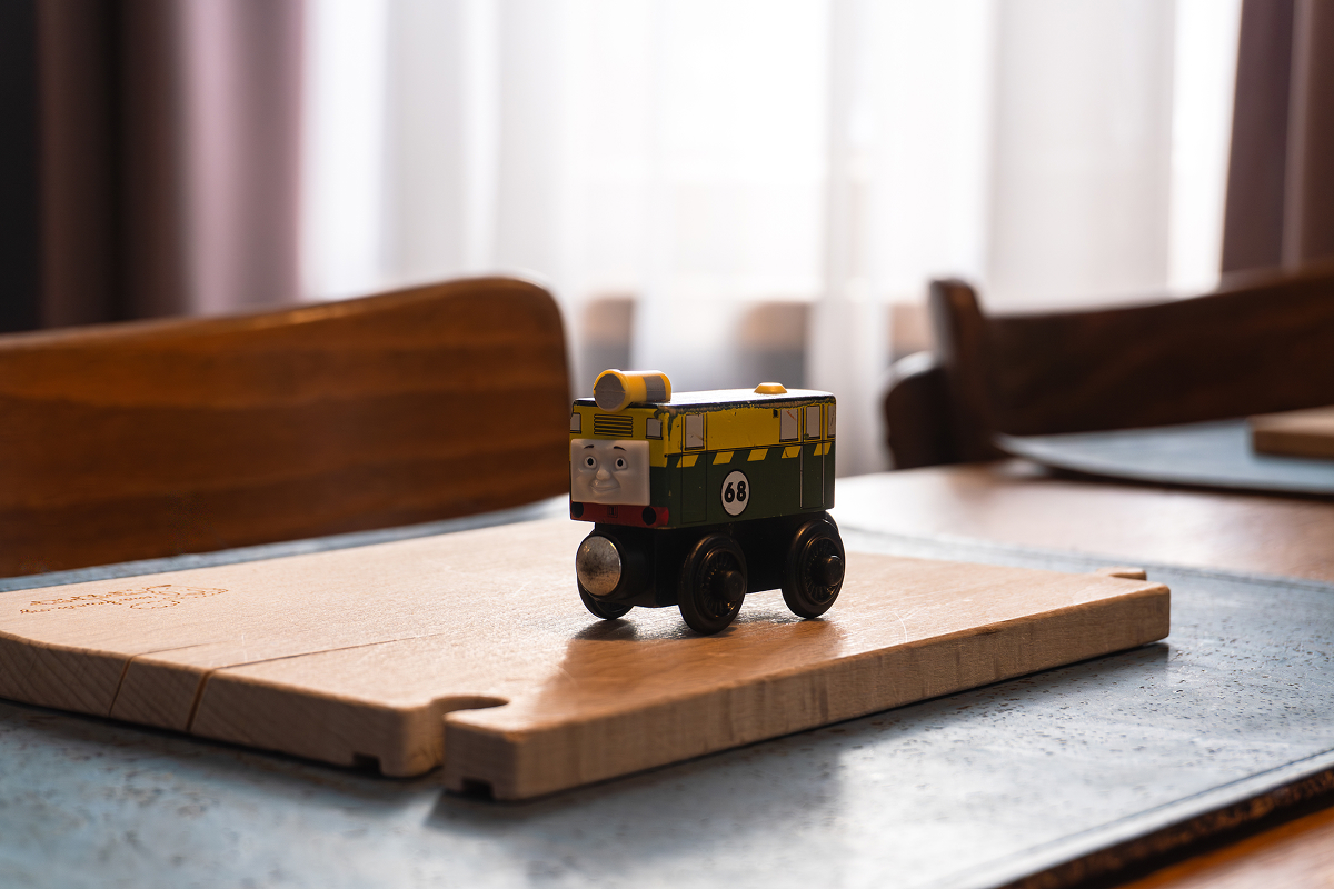 Eine kleine Spielzeugeisenbahn mit einem Gesicht und der Zahl 68 sitzt auf einem Holzbrett auf einem Tisch, mit verschwommenen Stühlen und einem hellen Fenster im Hintergrund.