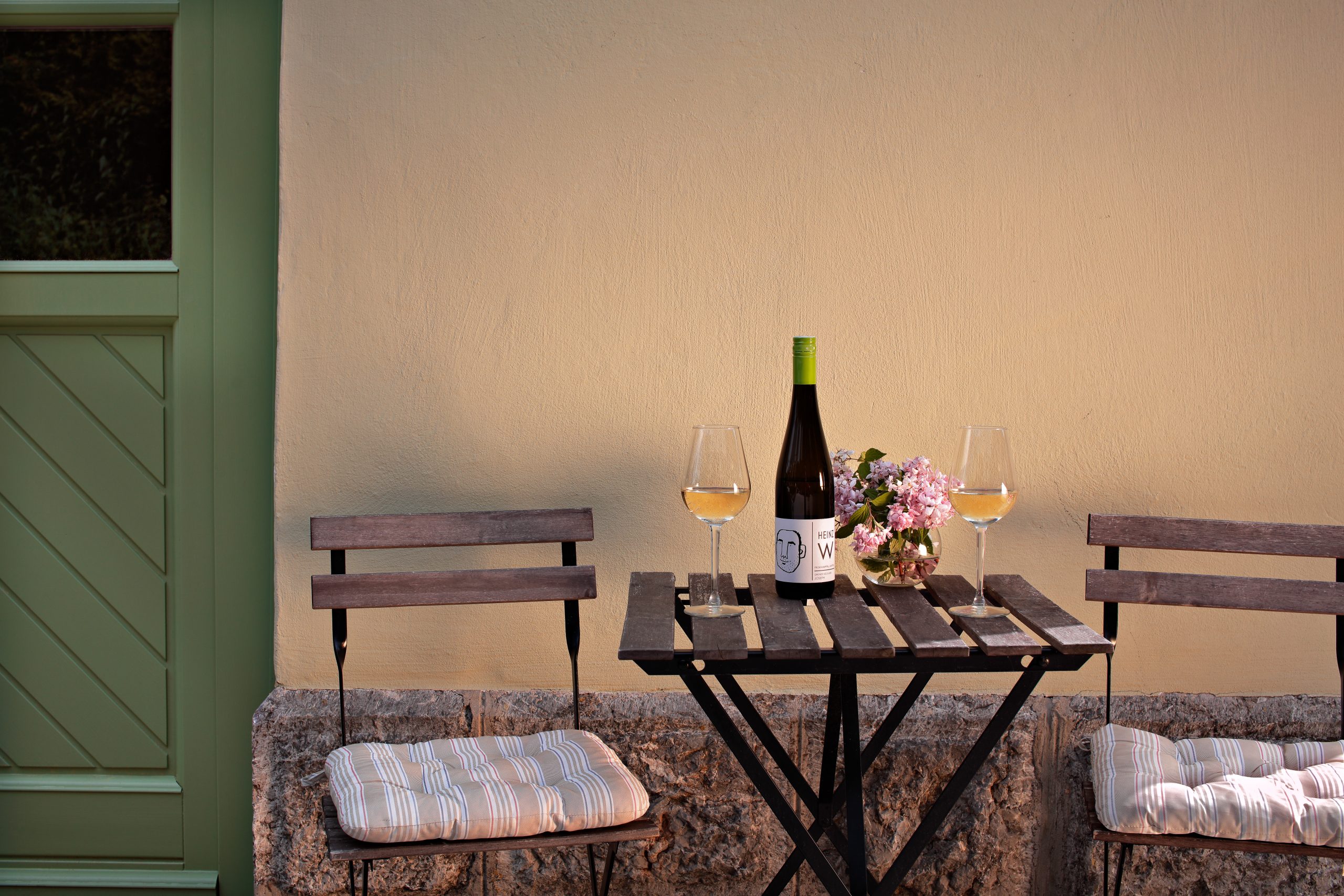 Ein kleiner Tisch mit zwei gepolsterten Stühlen im Freien steht mit einer Weinflasche, zwei Gläsern Weißwein und einer kleinen Vase mit rosafarbenen Blumen vor einer beigen Wand und neben einer grünen Tür.