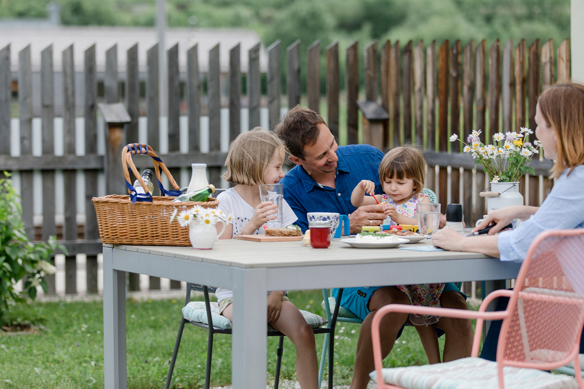Eine vierköpfige Familie sitzt an einem Picknicktisch im Freien und genießt eine gemeinsame Mahlzeit. Auf dem Tisch steht ein Picknickkorb und im Hintergrund ist ein Holzzaun zu sehen. Die Atmosphäre ist entspannt und fröhlich.