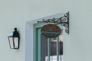 Ein dekoratives Metallschild mit der Aufschrift Zur Himmelsstreppe hängt neben einer Leuchte an einer hellen Wand über einem Fenster mit senkrechten grünen Balken.