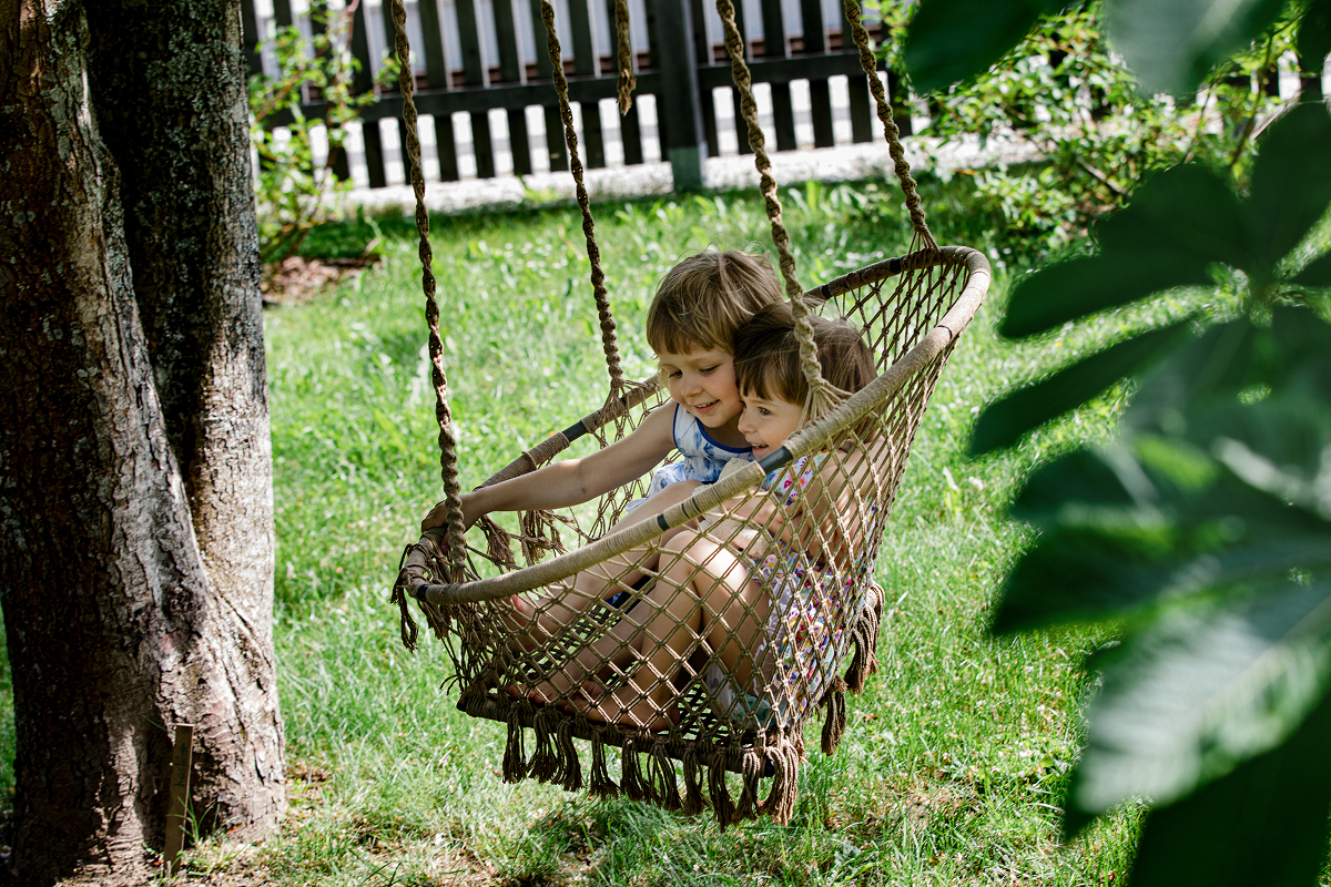 Zwei kleine Kinder sitzen eng beieinander in einer geflochtenen Hängeschaukel, lächeln und genießen einen sonnigen Tag in einem grünen Garten, mit Bäumen und einem Zaun im Hintergrund.
