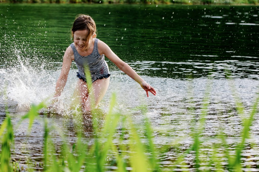 Ein junges Mädchen in einem gestreiften Badeanzug plantscht und spielt an einem sonnigen Tag vergnügt in einem flachen See, umgeben von grünem Gras und Wasserpflanzen.