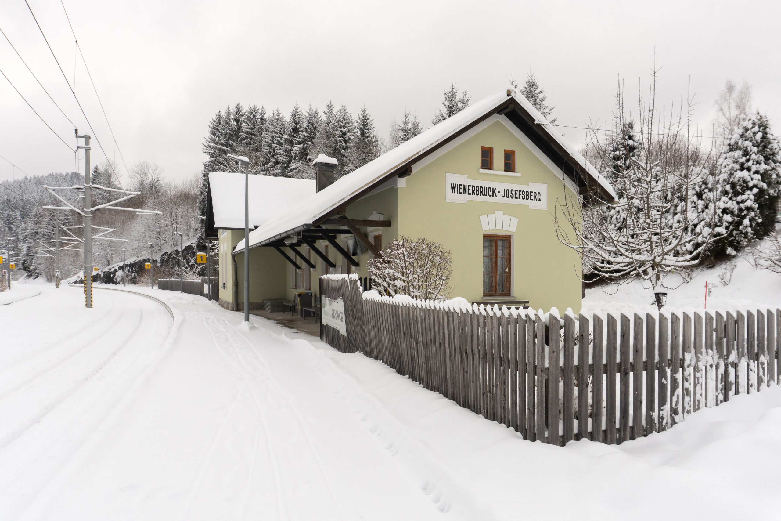 urlaubambahnhof-winter-ariane-kabengi-DSC3211 Ein kleines Bahnhofsgebäude mit einem Schild mit der Aufschrift Weberbruck-Josefsberg steht neben schneebedeckten Gleisen und einem Holzzaun, umgeben von Bäumen und einer frisch verschneiten Landschaft.