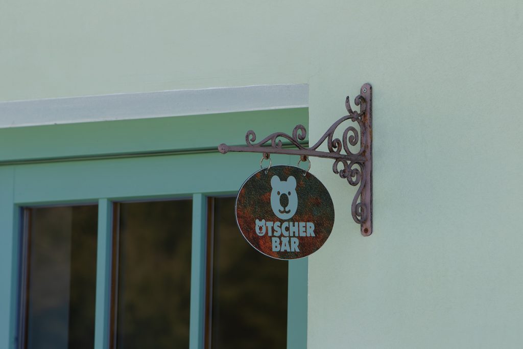 Ein dekoratives Metallschild mit einem Bärengesicht und dem Schriftzug Ötscher Bär hängt an einer verzierten Halterung vor einem pastellgrünen Gebäude mit einem türkisfarbenen Fensterrahmen.