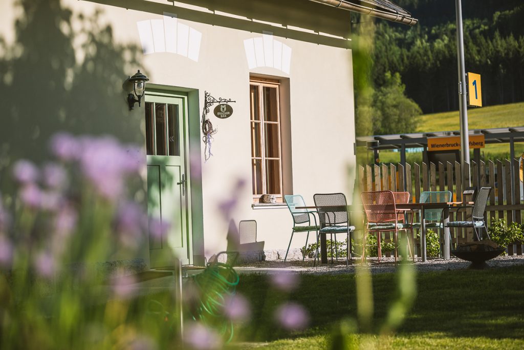 Ein charmanter Sitzbereich im Freien mit bunten Stühlen und Tischen neben einem hellgelben Gebäude, umgeben von Grün und Blumen, mit einem Zaun und einem gelben Schild im Hintergrund.