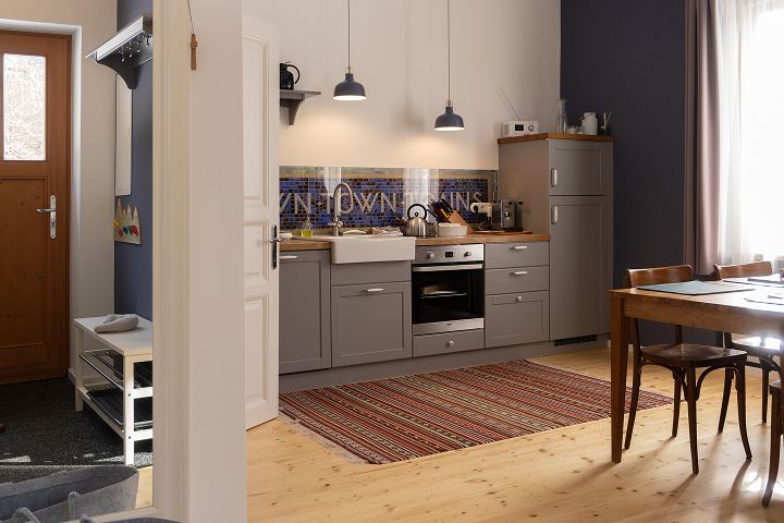 Eine gemütliche Küche mit grauen Schränken, einer gefliesten Aufkantung, Geräten aus Edelstahl und Hängeleuchten. Ein gestreifter Teppich bedeckt den Holzboden, und ein hölzerner Esstisch mit Stühlen steht an einem sonnendurchfluteten Fenster.