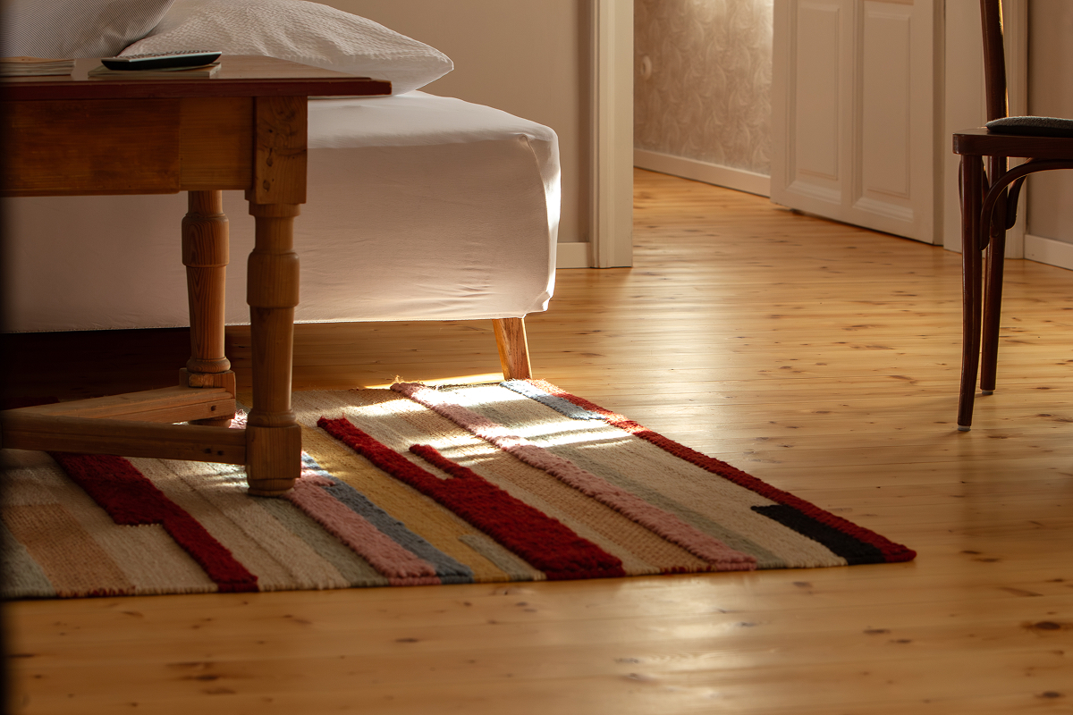 Ein bunt gestreifter Teppich liegt auf dem Holzboden neben einem Bett mit weißem Bettzeug. Sonnenlicht strömt durch eine offene Tür herein und wirft warmes Licht in den Raum. Ein Holzstuhl und ein Tisch sind teilweise sichtbar.