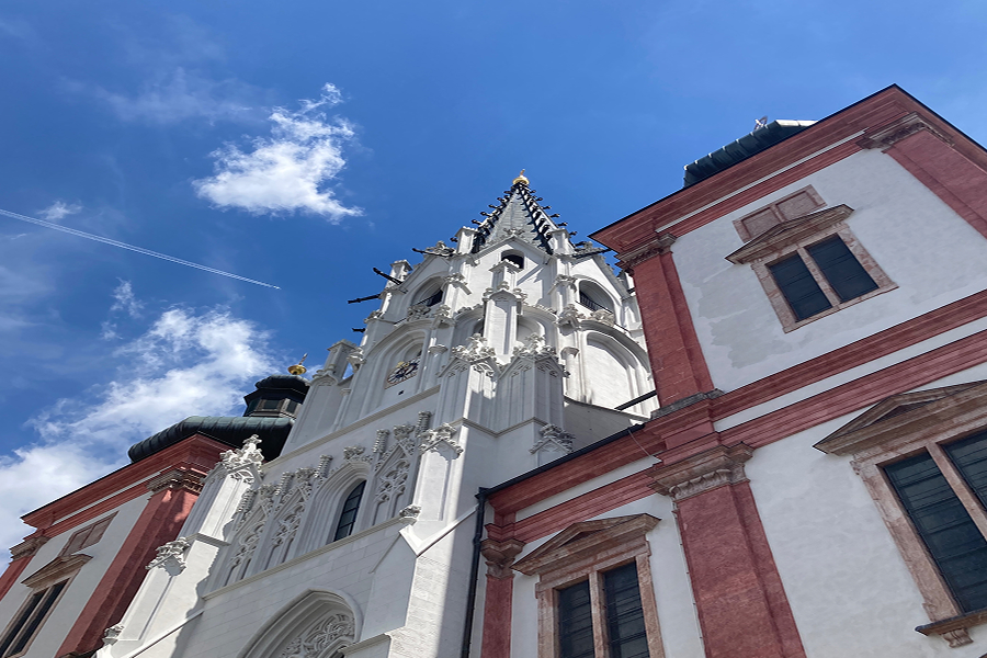 mariazell-basilika-IMG_0478 Eine hohe, verschnörkelte Kirche mit weißen und rosaroten Verzierungen steht vor einem strahlend blauen Himmel mit Wolkenfetzen, gesehen aus einem niedrigen Winkel. Ein Flugzeug hinterlässt einen Kondensstreifen am Himmel über der Kirchturmspitze.