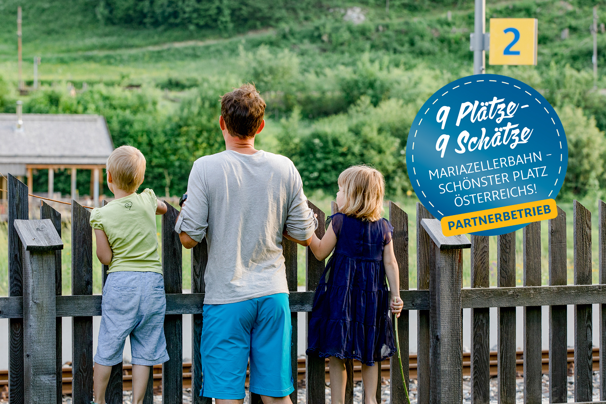Ein Mann und zwei kleine Kinder stehen an einem Holzzaun und blicken auf die Bahngleise, im Hintergrund erstreckt sich eine üppig grüne Landschaft. Ein rundes blaues Schild in deutscher Sprache wirbt für die Mariazellerbahn als Österreichs schönsten Ort.