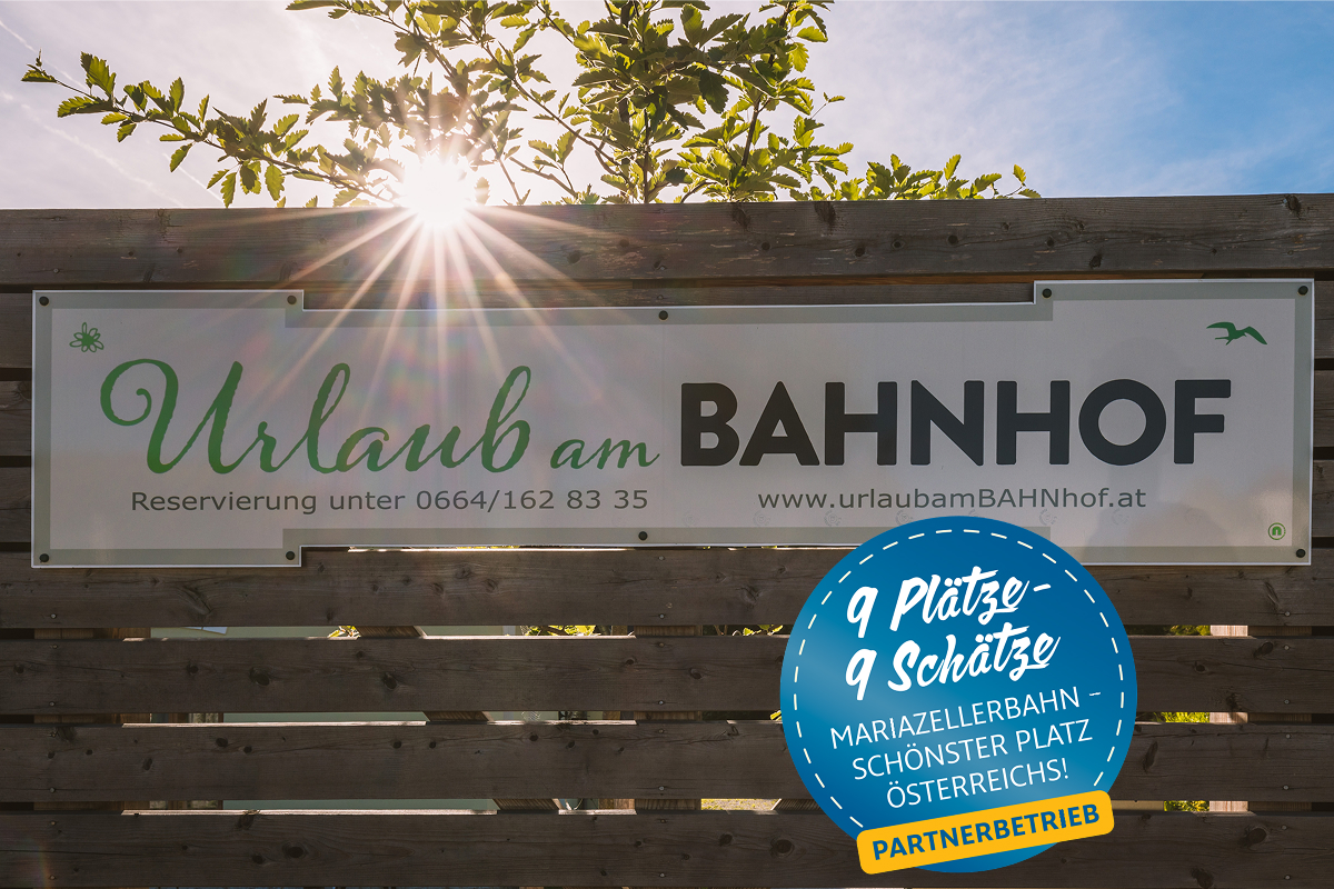 Auf einem Banner an einem Holzzaun steht „Urlaub am BAHNHOF“, darunter sind die Kontaktdaten angegeben. Auf einem blauen Schild steht: „9 Plätze – 9 Schätze, Mariazellerbahn – schönster Ort Österreichs! Partnerbetrieb.“ Hinter dem Zaun scheint die Sonne.